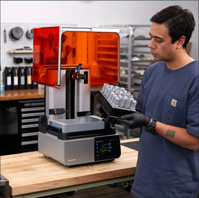 a build plate loaded with multiple small 3D printed parts next to a modern resin 3D printer with an orange UV cover