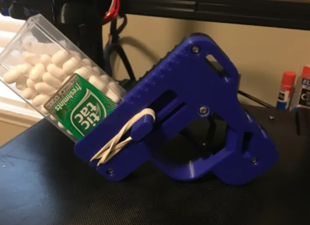 A blue 3D-printed Tic Tac Gun V2 Rubber Band Edition loaded with a box of white Tic Tacs and powered by a rubber band.