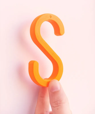 A hand holding a bright orange, smoothly finished 3D printed S-hook against a light pastel background.