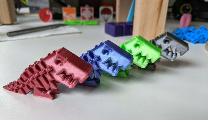 A colorful lineup of 3D-printed articulated Flexi Rex dinosaur toys in red, blue, green, and grey, sitting on a white desk