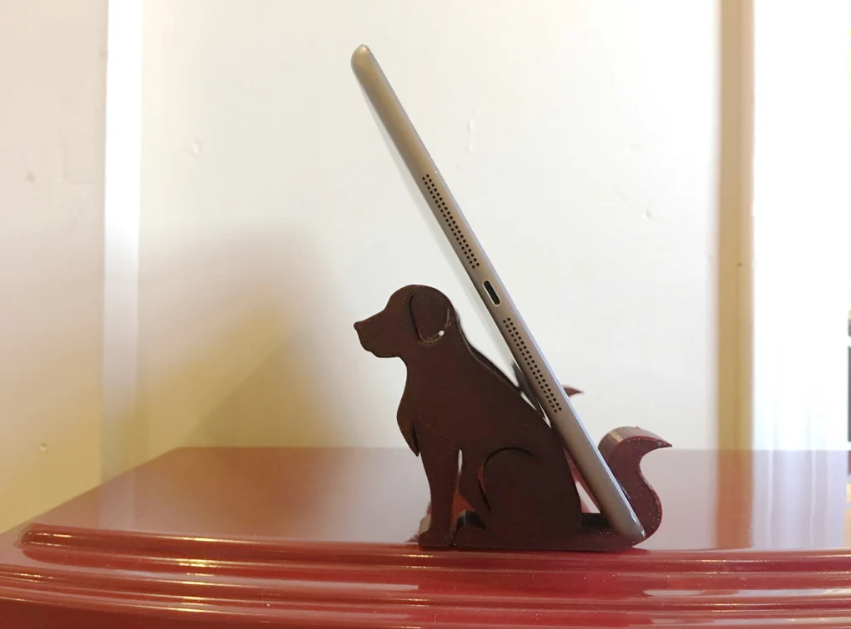 A silhouette-style 3D-printed stand shaped like a sitting dog, supporting a tablet against its back on a red surface.