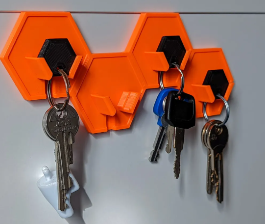 A bright orange 3D-printed wall key hanger made of interlocking hexagonal tiles, with black inserts holding keychains.