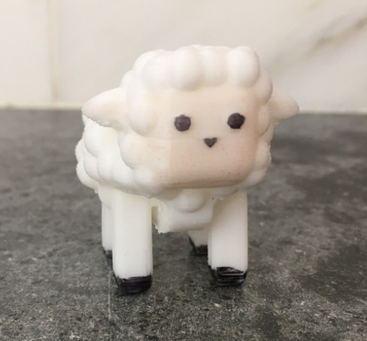 A cute, small 3D-printed sheep figurine with a bumpy white wool texture, black legs, and simple black facial features, standing on a dark countertop