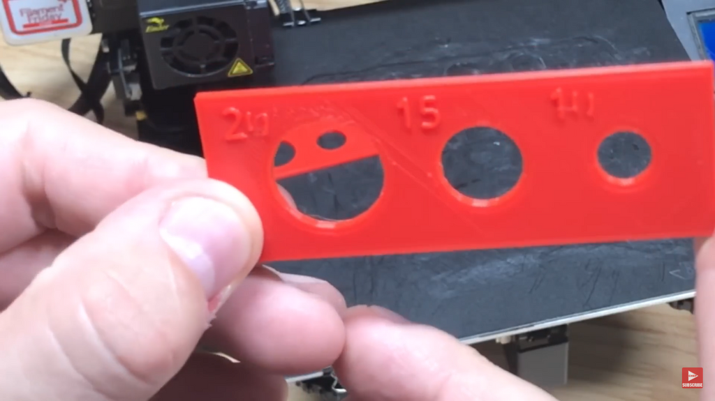 A finished red 3D printed test part held up to the camera, showing three holes of different sizes (20mm, 15mm, 10mm) after successful horizontal expansion calibration.