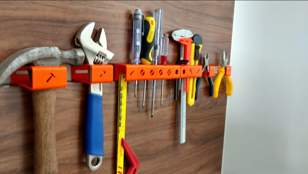 A bright orange, wall-mounted 3D-printed tool and hex bit holder keeping screwdrivers, wrenches, pliers, and a hammer neatly organized on a wooden backboard.