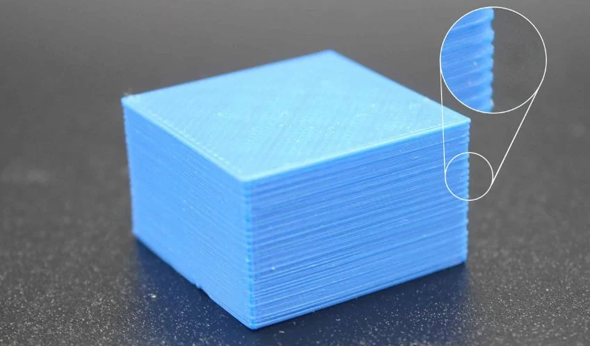 A blue 3D printed cube demonstrating over-extrusion, with a magnified inset showing excessive filament causing messy, bulging layers.