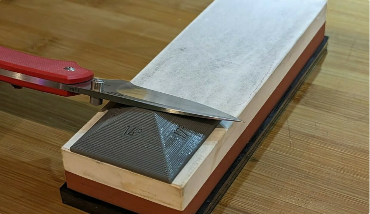 A grey 3D-printed angle guide block resting directly on a two-toned whetstone, holding a kitchen knife blade at the perfect angle for sharpening.