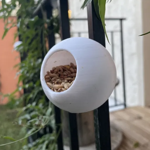 A white, spherical 3D printed bird feeder filled with seeds, hanging outdoors near a balcony.