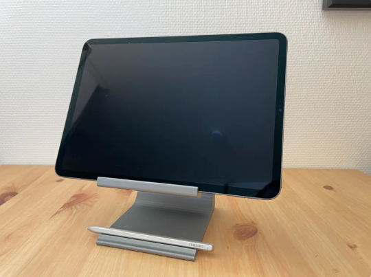 A sleek, silver-colored 3D-printed desk stand with a wide base and a dedicated slot for an Apple Pencil, holding a large tablet.