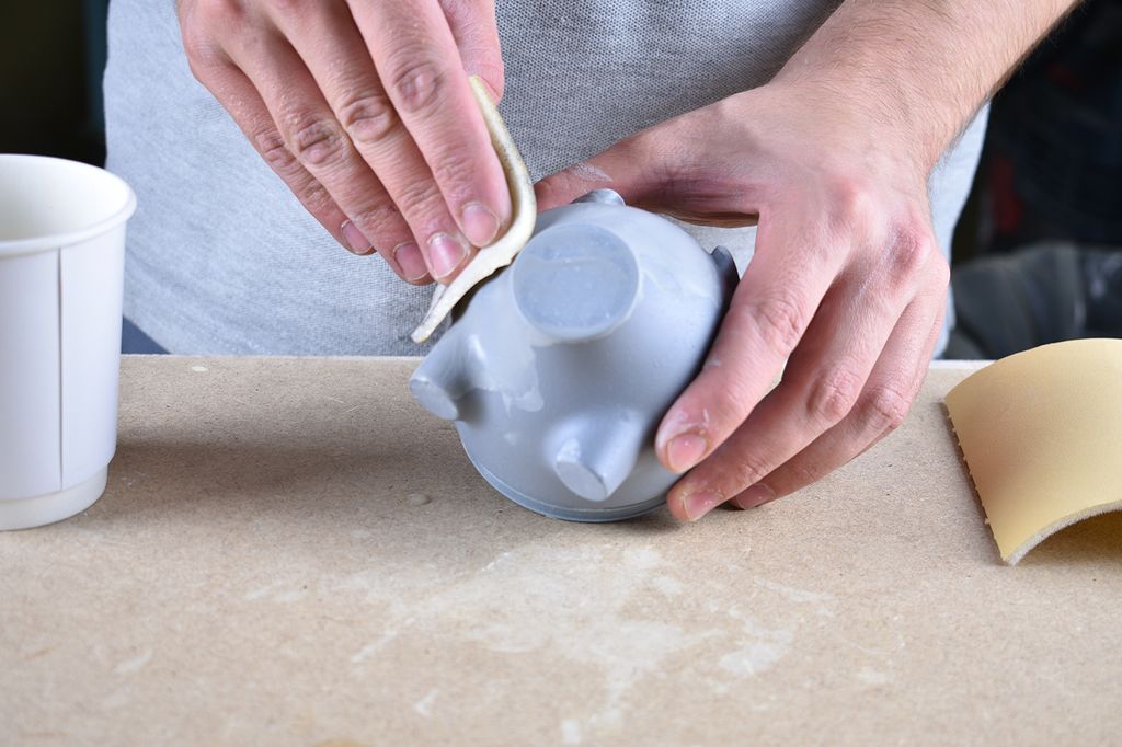 Hands using a piece of sandpaper to smooth the surface of a grey 3D-printed piggy bank.  