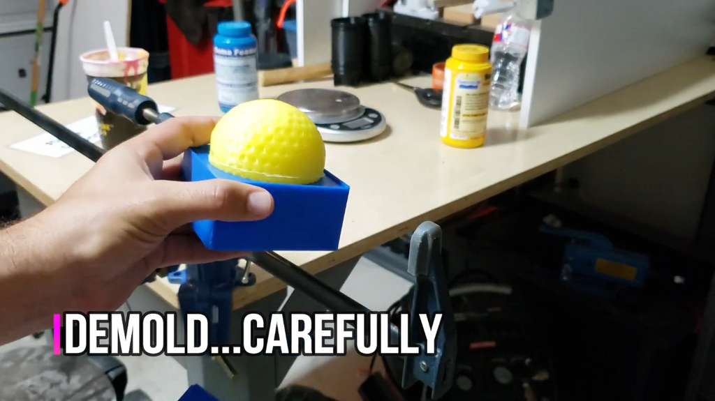 A hand carefully pulling a cast yellow textured ball out of a blue 3D-printed mold base on a workbench.