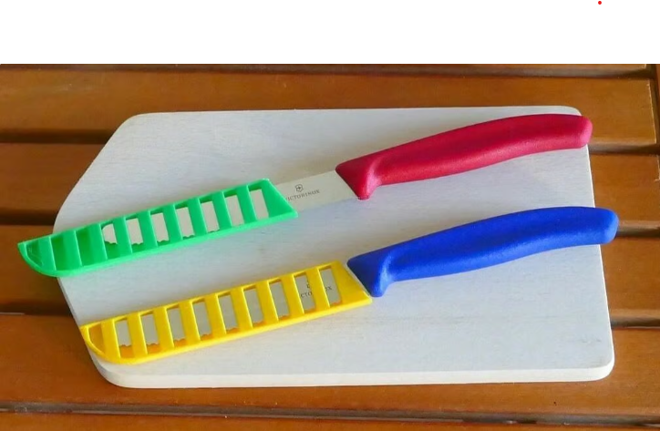 Two kitchen knives placed on a white cutting board, their blades protected by brightly colored, slotted 3D-printed sheaths in green and yellow.