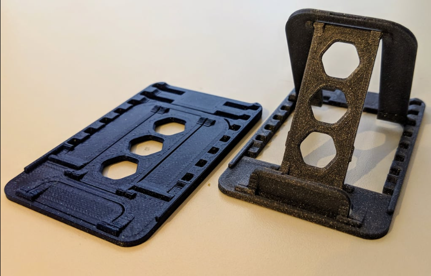 Two black 3D-printed portable stands; one is folded flat like a credit card, and the other is unfolded to show the adjustable support frame.