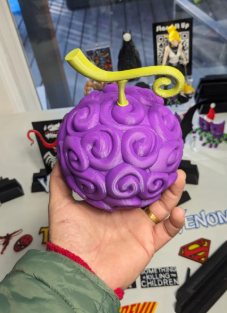 A person's hand holding a 3D printed replica of the purple Gomu Gomu no Mi Devil Fruit, featuring its signature swirl patterns and a yellow stem