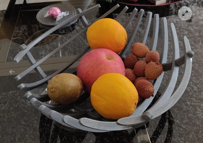 A grey, modern 3D-printed fruit bowl featuring a curved, slatted design, filled with an apple, oranges, kiwi, and lychees