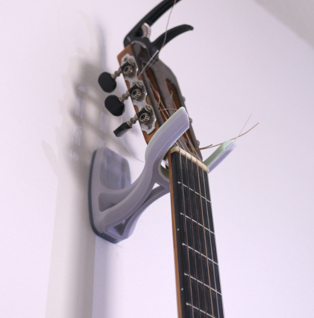 A grey and translucent 3D printed guitar wall mount hanger securely holding the neck of an acoustic guitar against a white wall