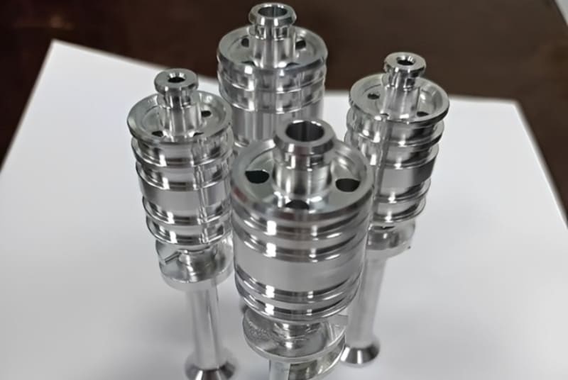 Four highly polished, precision-machined cylindrical metal parts standing upright on a white surface.