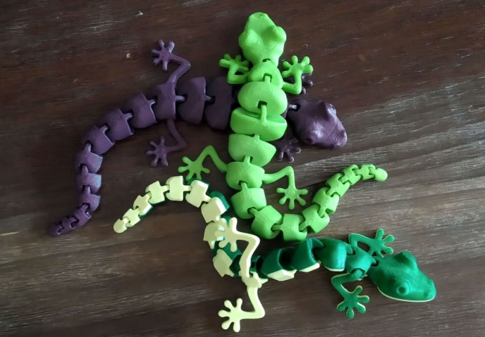 Three 3D-printed articulated lizards in green, light green, and purple, featuring realistic print-in-place segmented joints resting on a wooden surface