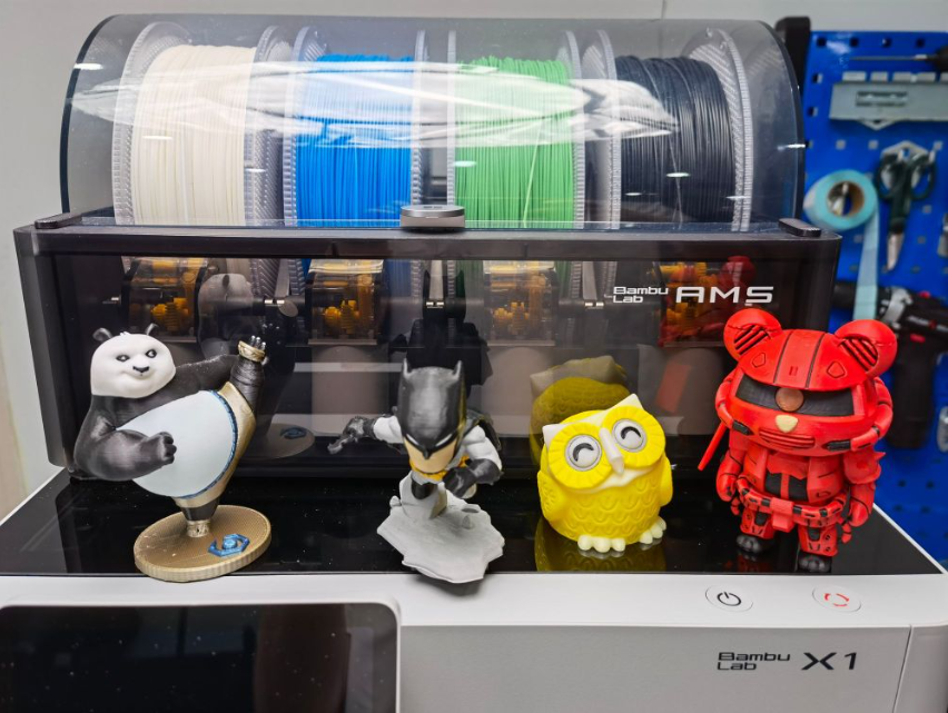 A Bambu Lab 3D printer equipped with the Automatic Material System (AMS), showcasing four different colored filament spools loaded into the top enclosure.