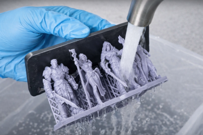 A blue-gloved hand rinsing a build plate loaded with 3D-printed fantasy miniatures under a running water ta