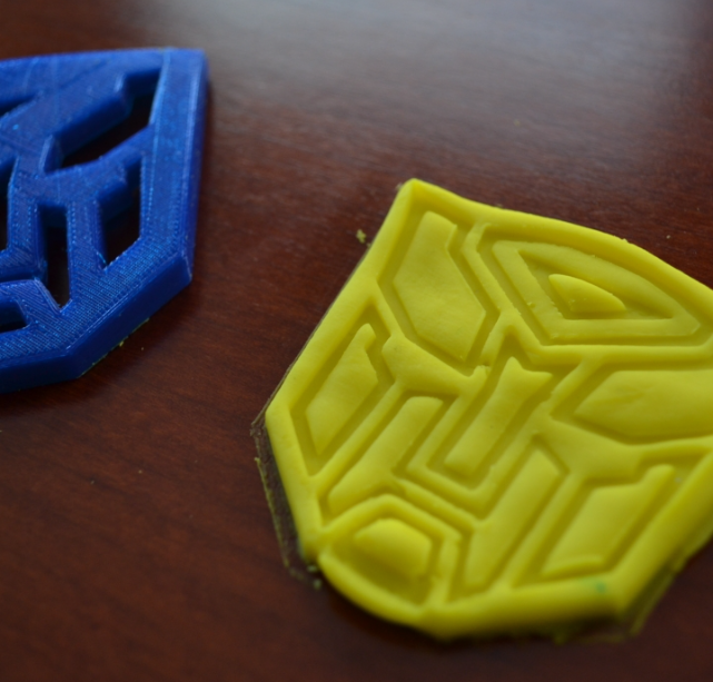 Two 3D printed Transformers cookie cutters, one blue Decepticon logo and one yellow Autobot logo, resting on a wooden table.