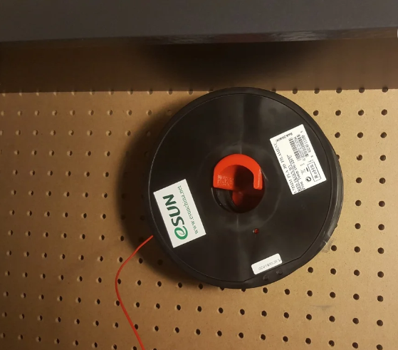 An orange 3D printed pegboard filament spool holder securely displaying a black eSUN filament spool on a wooden pegboard wall.