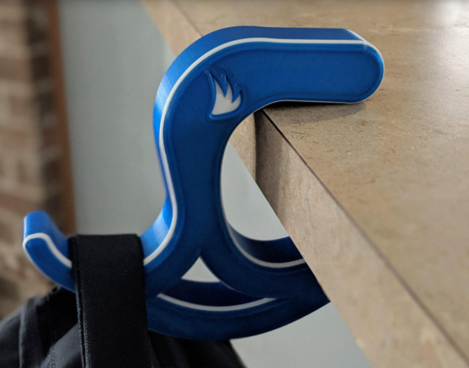 A blue and white 3D printed desk hook known as "The Claw," firmly attached to the edge of a desk and holding a heavy black backpack strap.