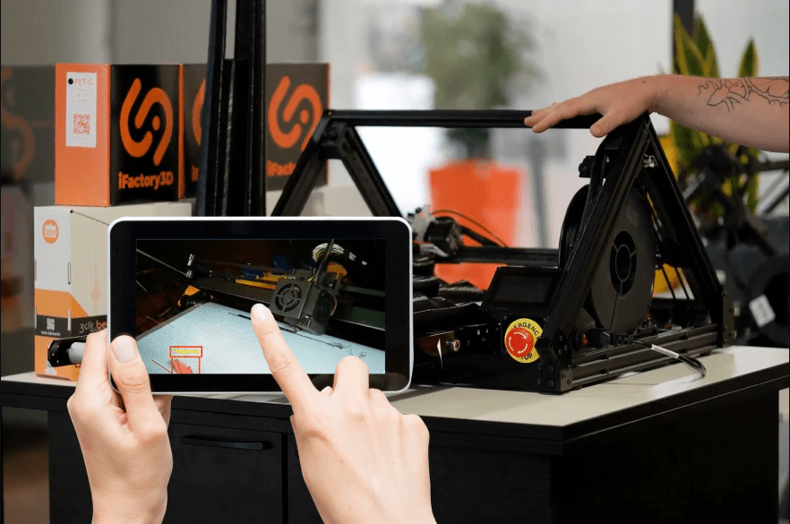 A user holding a tablet displaying the iFactory One AI camera feed detecting an error on the print bed.  