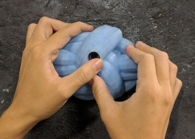 A pair of hands firmly gripping a light blue, bulbous, 3D-printed VR climbing hold mounted on a dark, textured wall.