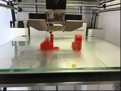 Two red 3D printed house models on a glass print bed, demonstrating poor bed adhesion where the base of the prints are warping and lifting from the surface.