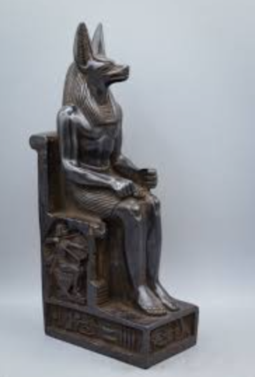 A weathered dark stone statue depicting the ancient Egyptian god Anubis seated on a throne.