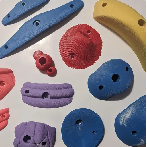 A vibrant assortment of 3D-printed climbing holds in blue, red, yellow, and purple, featuring various shapes like jugs, crimps, and pinches, laid out on a white surface.