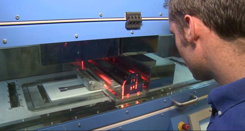 A technician monitoring a High-Speed Sintering (HSS) 3D printer in action, showing the infrared lamp moving across the polymer powder bed.