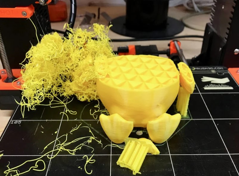 A botched 3D print with yellow plastic spaghetti waste on the printer bed next to a partially formed object.