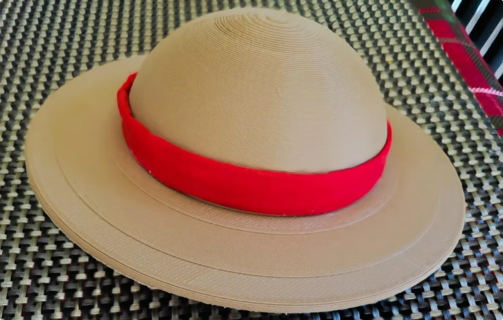 A realistic 3D printed replica of Luffy's iconic straw hat printed in tan filament and wrapped with a bright red fabric ribbon.