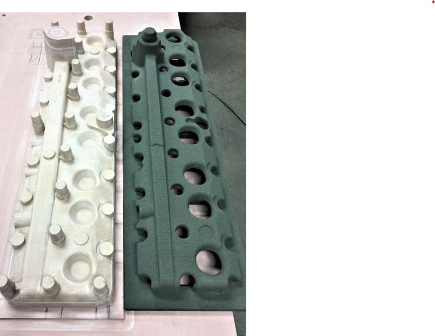 A large, complex light beige 3D-printed sand mold sitting next to its corresponding dark green metal cast part.