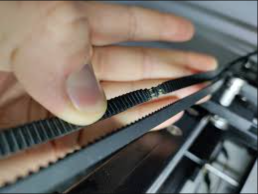 A close-up view of a finger lifting a loose black timing belt on an FDM 3D printer to inspect its tension.