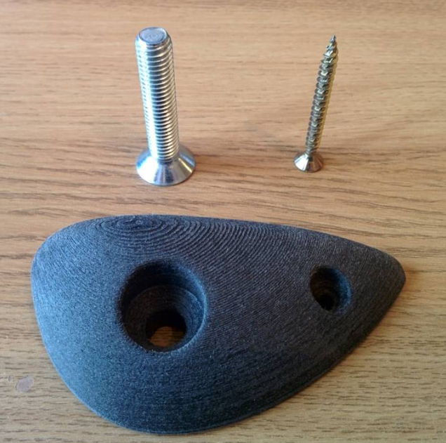 A dark grey, textured, 3D-printed climbing hold placed on a wooden surface next to a large countersunk bolt and a smaller wood screw.