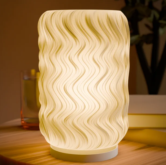 A glowing, cylindrical 3D printed lampshade with wavy, undulating vertical lines reminiscent of a tide pattern, resting on a table