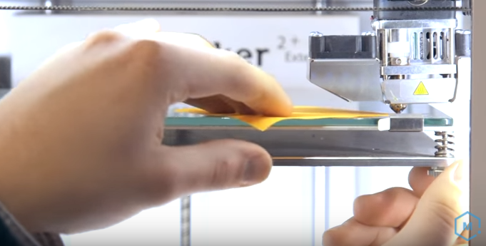 A person using a piece of paper as a feeler gauge to manually adjust the thumb screws and level a 3D printer bed.