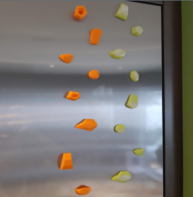 Multiple miniature climbing holds in bright orange, yellow, and green attached as magnets to the surface of a stainless steel refrigerator.
