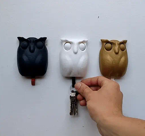 Three 3D printed owl wall key holders in black, white, and gold, with a hand demonstrating how to insert the interchangeable printed eyes.