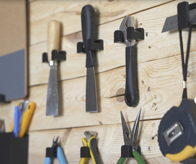 A collection of functional black 3D printed tool hooks mounted on a wooden board, neatly organizing workshop tools like chisels, pliers, and a tape measure.