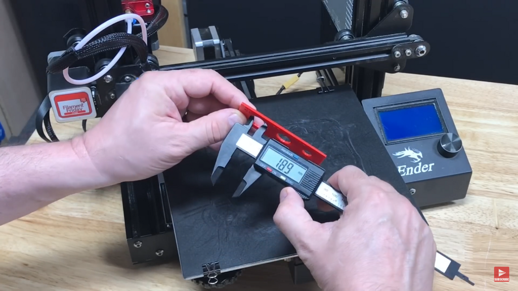 A person using digital calipers to measure the diameter of a hole in a red 3D printed test part to check for dimensional accuracy against the original design.