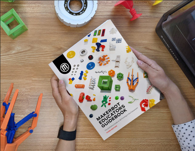 Top-down view of a teacher holding the MakerBot Educator's Guidebook on a wooden desk surrounded by colorful 3D printed objects and a 3D printer.