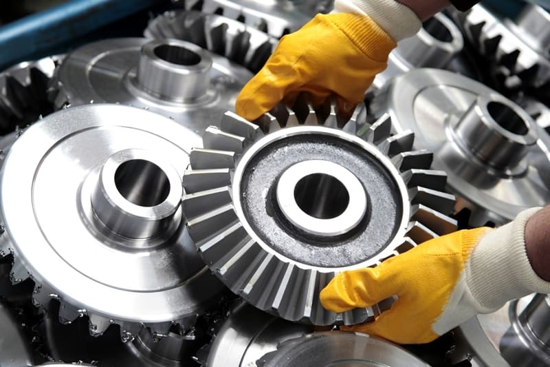 A pair of yellow-gloved hands holding and carefully inspecting large, shiny, precision-machined metal gears.