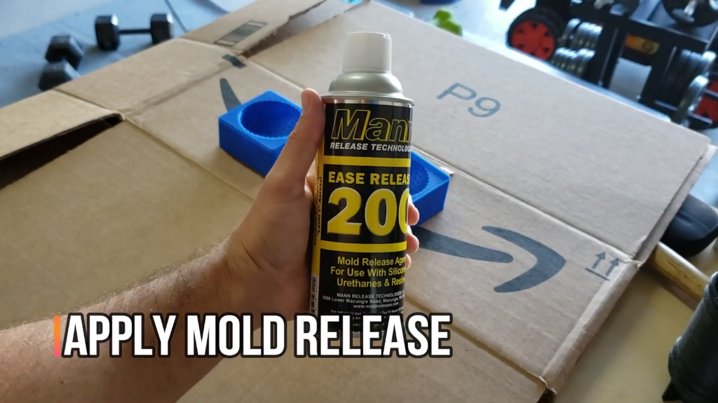 A hand holding a can of Mann Ease Release 200 spray over a cardboard work surface, ready to prep a blue 3D-printed mold.