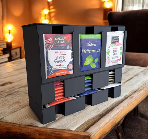 A sleek, grey 3D-printed tea bag dispenser with three slotted sections displaying different colorful tea bags, resting on a wooden tabletop