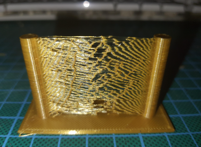 A golden 3D print showing heavy stringing and oozing artifacts between two vertical posts.  