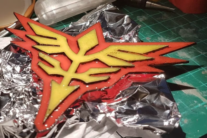 A dual-extrusion 3D-printed Neo Zeon logo featuring a yellow emblem with a red border resting on aluminum foil.